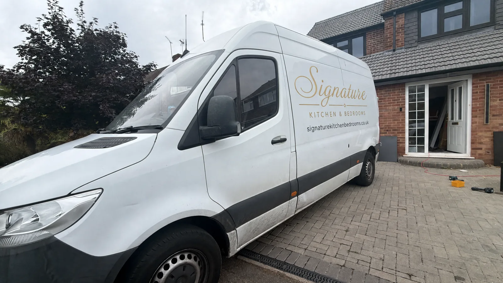 A white Mercedes Sprinter van with Signature Kitchen and Bedrooms lettered in gold on the side, parked on a block-paved driveway outside a house with the front door open and tools on the ground