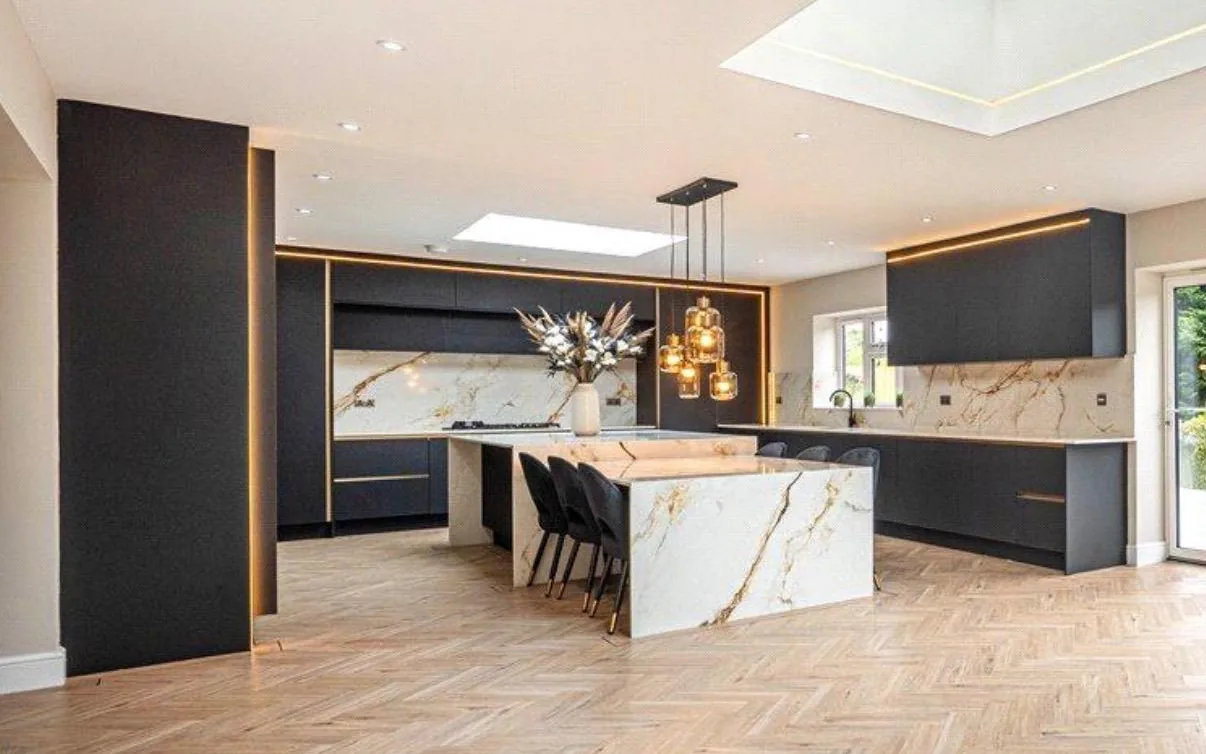 An open-plan fitted kitchen in matt navy with a gold-veined marble island and splashback, brass trim and a herringbone oak floor