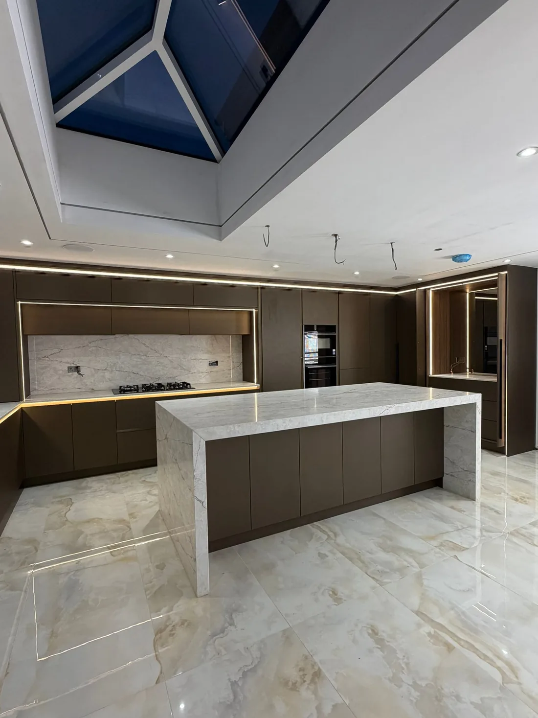 The island seen from the hob side: six matt bronze doors under a veined white worktop that runs down both ends to the polished floor, with the hob recess and its veined splashback behind