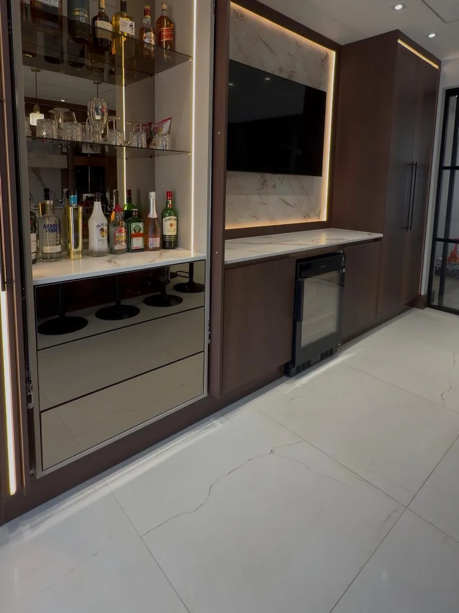A bespoke home bar in dark walnut with a mirrored back panel, lit glass shelving, a stone worktop and an integrated wine fridge