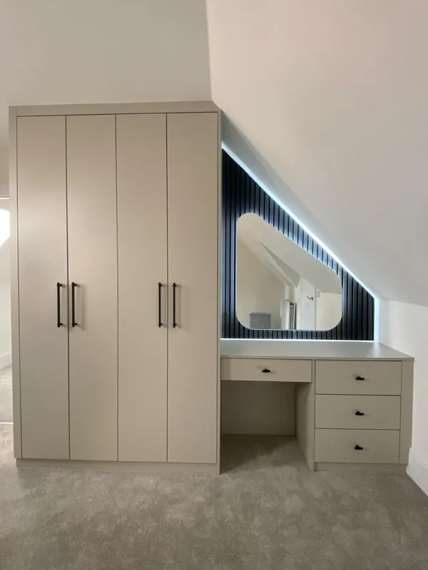 A loft bedroom with cream fitted wardrobes and a dressing table built into the eaves, behind a navy fluted panel with concealed lighting