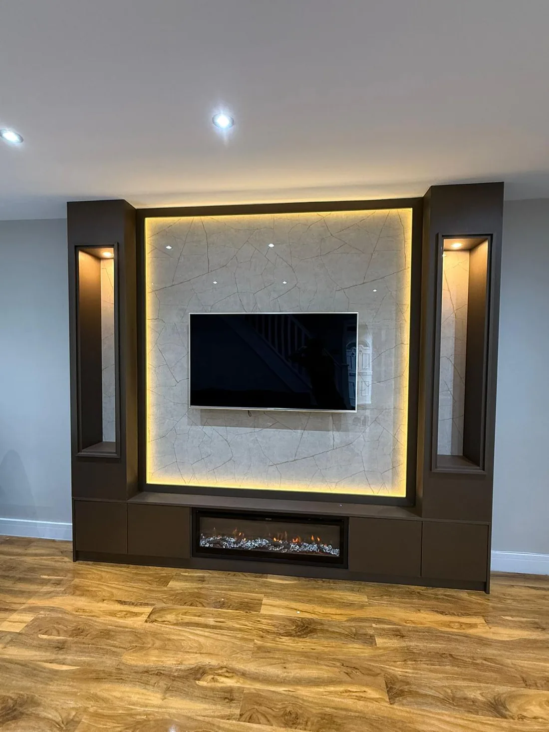 A media wall in bronze-brown panelling with a backlit stone centre panel, a flush television, a linear electric fire and lit display recesses