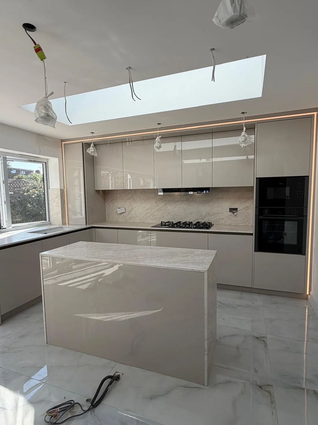 A cream gloss handleless kitchen with a marble worktop and island, integrated ovens and a full-length skylight above